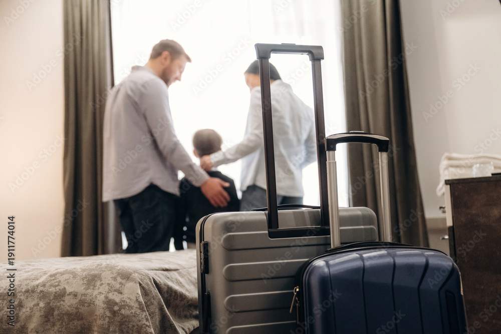 Fototapeta premium Family of three standing together in a hotel room with luggage on the bed, large windows and curtains visible in the background creating a cozy atmosphere