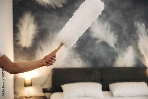 Картината върху платно Hand holding a white feather duster cleaning a hotel room with a modern bed and