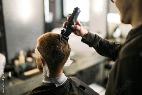 Photos Caucasian young adult man sitting in barbershop chair receiving haircut from Cau