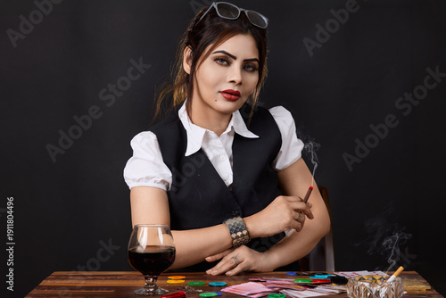 Portrait of a confident woman seated at a gambling table holding a cigarette, surrounded by poker chips, cards and alcohol. Concept image illustrating power, high risk decisions, casino lifestyle