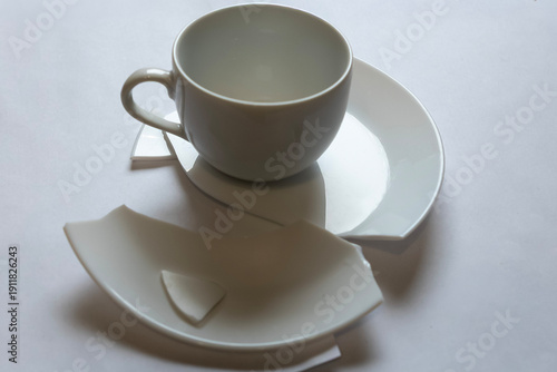Broken cup and plate are placed on a simple background showing fragments and pieces of white pottery scattered around