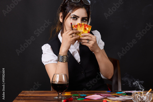 Portrait of a confident woman holding playing cards in front of her face while smoking at a gambling table surrounded by poker chips and alcohol. Concept image representing strategy, secrecy