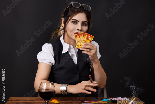 Portrait of a confident woman holding playing cards while smoking at a gambling table surrounded by poker chips and alcohol. Concept image illustrating strategy, competition, high risk decisions