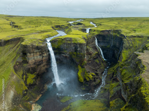 Aerial view of the cascading waters of Haifoss waterfall plunge into the gorge below, surrounded by mossy green cliffs under a cloudy sky, Haifoss, Iceland.