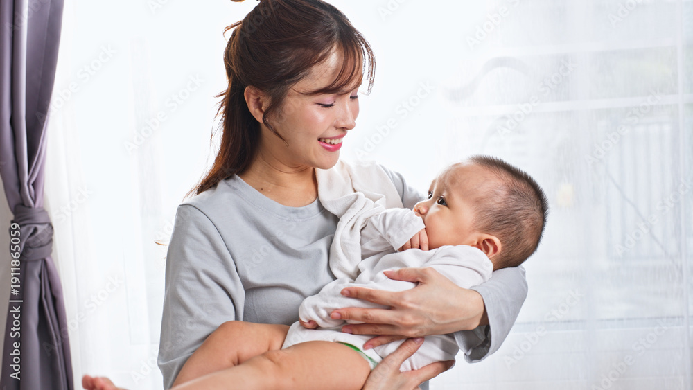 Obraz premium Young happy asian mother holding adorable infant baby near window in bright room showing love care motherhood tenderness and family bond in domestic lifestyle