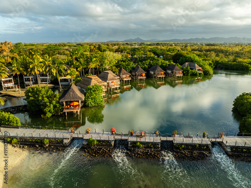 Aerial view of thatched-roof bungalows standing above the water, reflecting the sky, surrounded by lush greenery, Prince Maurice, Flacq District, Mauritius.
