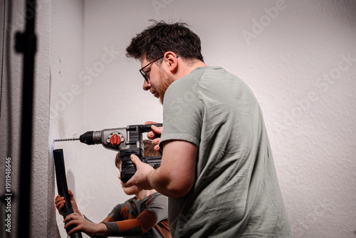 Indoor DIY renovation moment: an adult drills into a wall while a child helps by holding a vacuum hose to capture dust. Real family teamwork for home improvement and installation.