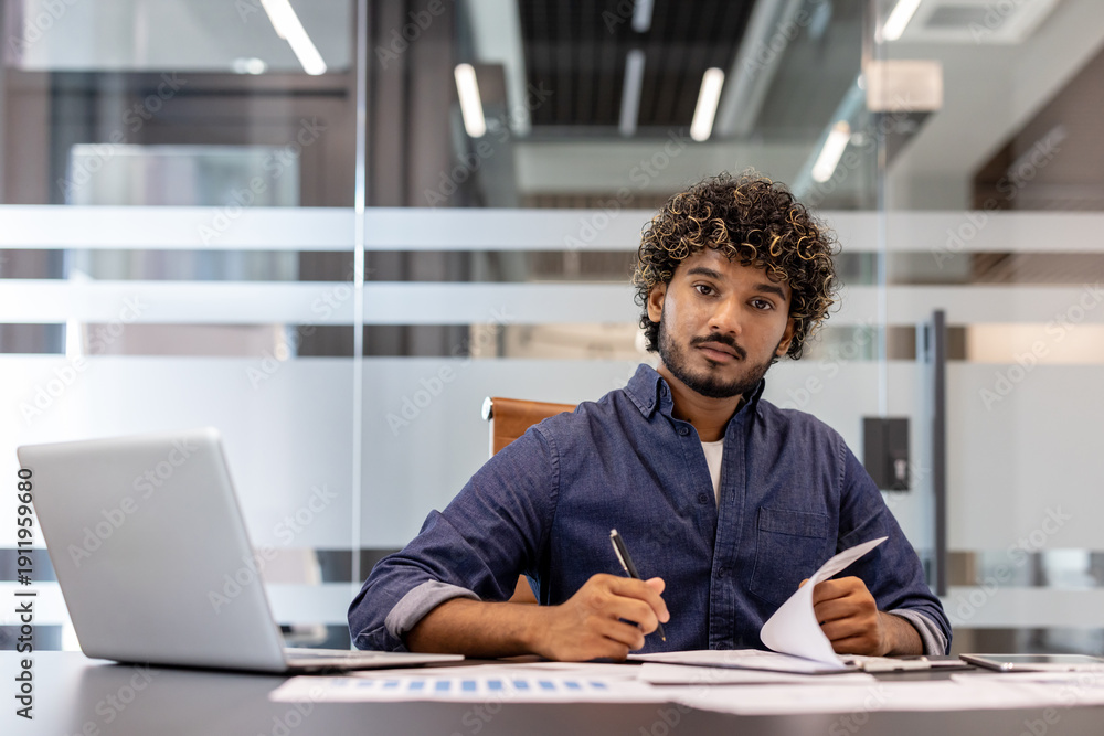 Fototapeta premium Portrait of a serious young Indian businessman working in the office with a laptop and documents, looking at the camera