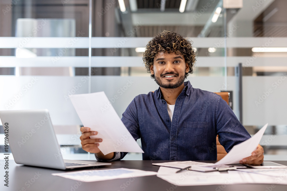 Fototapeta premium Portrait of a smiling young Indian businessman sitting in the office in front of the camera, working on a laptop and with documents