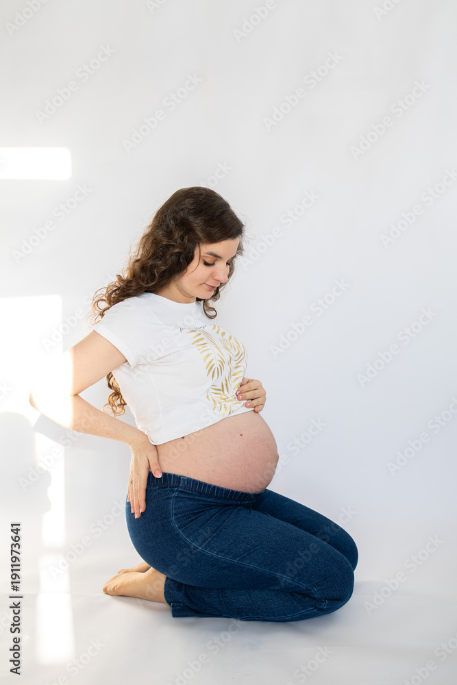 Fototapeta premium Beautiful pregnant woman in white t shirt and jeans posing in studio. Maternity tenderness parenthood new life. Happy pregnant woman on white background.