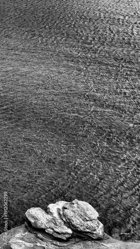 Black and white minimalist coastal scene featuring layered rock formations in the foreground and wind-rippled seawater creating textured patterns across the surface. The high-contrast composition emph