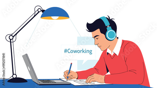 Young man with headphones writing notes and working on a laptop at a professional coworking space under a desk lamp.