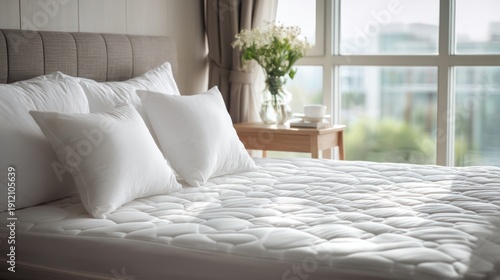 A neatly made bed with white pillows and a quilted mattress, with a window in the background showing a cityscape.