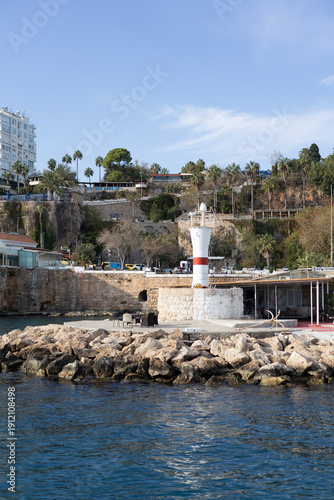 Small white lighthouse with a red stripe located on a stone pier near the coastline. Rocky shore with palm trees, buildings and coastal infrastructure in the background. 