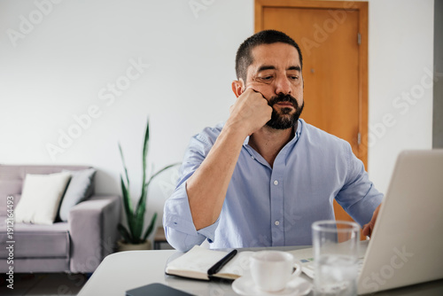 Obraz na płótnie Man sitting at desk resting face on hand while looking bored at laptop screen during slow work moment in home workspace with coffee and laptop