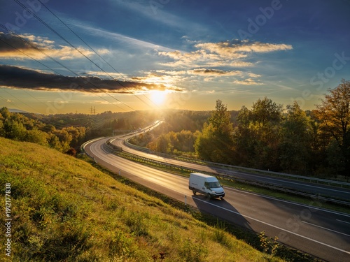 White delivery van driving on a winding highway through an autumn landscape at sunset