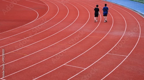 Two people are walking on a track. The track is red and has white lines. The people are walking in opposite directions