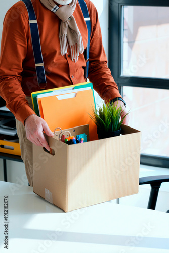 Adult man carrying cardboard box filled with office supplies and folders, standing near window in workspace, only torso and hands