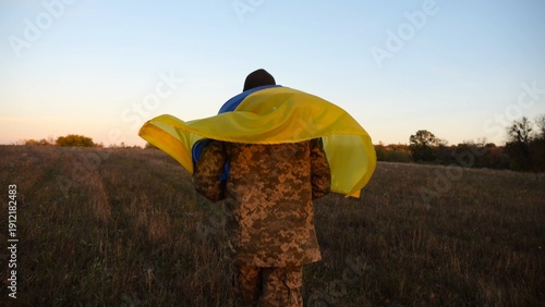 Young male military in uniform jogging with blue-yellow banner on shoulders at meadow. Soldier of ukrainian army running with flag of Ukraine as symbol of victory against russian aggression. Slow mo