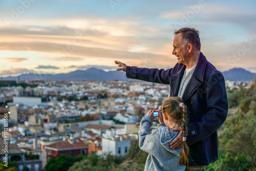 Father and his young daughter learning photography together during a walk, with Santa Bárbara mountain in Spain in the background, family bonding and creative lifestyle.