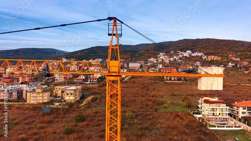 Tower crane counterweight and cabin with town background. Side view of a yellow tower crane showing the counterweight, cabin, and mast against a hillside town.