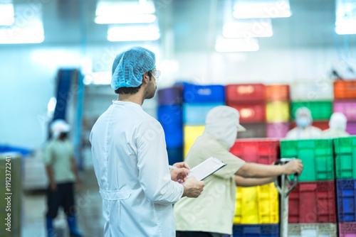 A male factory inspector in a lab coat, hairnet, and safety glasses stands and observes a busy food processing plant. Other workers move colorful crates in the background.