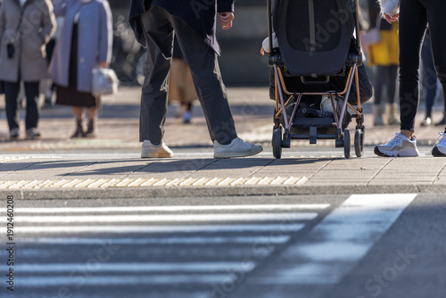 Photos Lower-angle of couple with child in stroller waiting on center median with pedestrians in bokeh background waiting to cross from opposite side