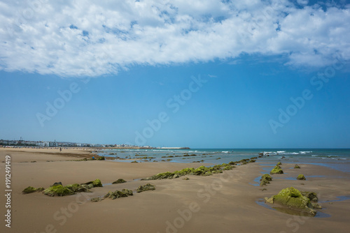 Wallpaper Mural Scenic beach in Asilah, Morocco with mossy rocks, calm ocean waves and a coastal town under a partly cloudy sky Torontodigital.ca