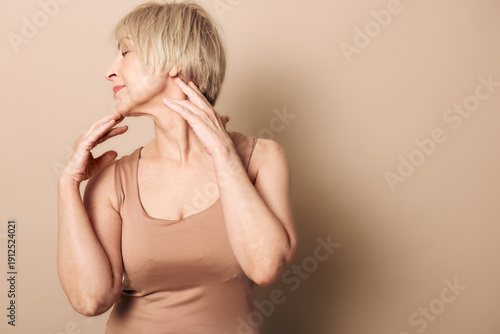 Fotografija Elegant mature woman in beige tank top poses gracefully with closed eyes and gentle smile against neutral background