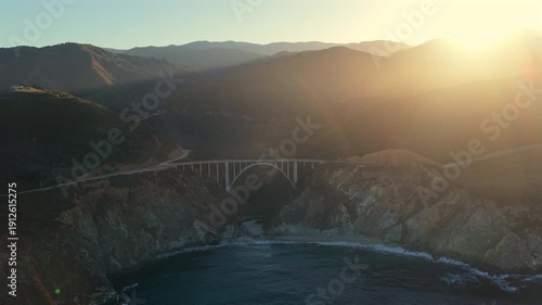 Aerial view of Bixby Creek Bridge in Big Sur California during golden hour, warm sunlight over coastal cliffs, Pacific Ocean waves, iconic highway and dramatic landscape