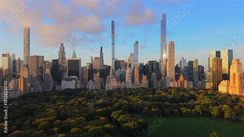 Aerial drone view of Manhattan skyline rising above Central Park in New York City, New York during golden hour with warm sunlight, long shadows and calm atmosphere.