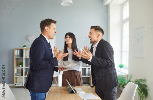 Fotografie In an office, two coworkers in argument at a desk while a colleague leads mediation and negotiation to resolve the dispute and reduce conflict