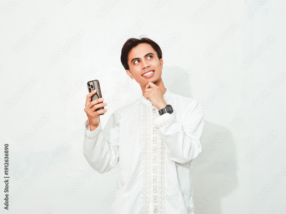 Fototapeta premium Asian Muslim man smiling while holding smartphone and looking up with thinking gesture, isolated on white background for technology and lifestyle advertising