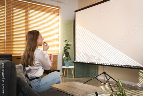 Young woman with popcorn and modern projector sitting on sofa in stylish livi...