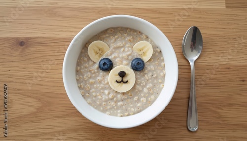 Cute bear face made with oatmeal, blueberries, and banana slices in a white bowl with a spoon on a wooden surface, a delightful and healthy breakfast
