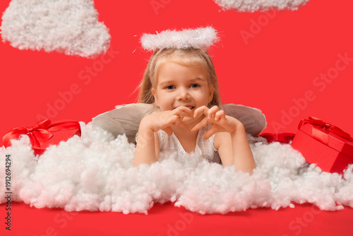 Cute little girl dressed as cupid with gift boxes and clouds showing heart gesture on red background. Valentine's Day celebration