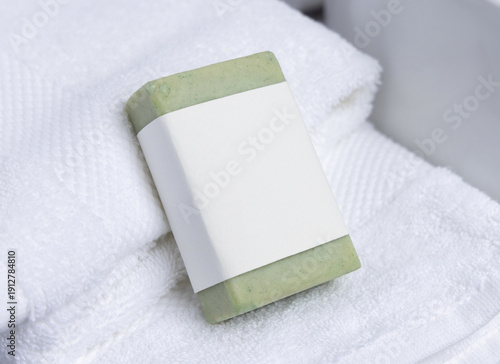 Green soap bar with blank label on white bath towels near basin, natural hygiene product mockup