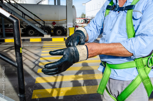 An industrial worker at a fuel loading terminal wearing safety gloves and a fall protection harness. A close-up of hands preparing for hazardous work. Workplace safety, personal protective equipment.