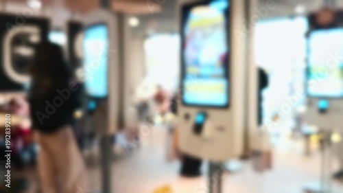 Unfocused footage of a modern fast-food restaurant interior. People use self-service digital kiosks with colorful food menus to order.