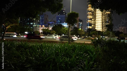 Nighttime scene featuring a cityscape with illuminated buildings and moving cars on a busy road, surrounded by greenery, showcasing the vibrant urban atmosphere and dynamic traffic flow