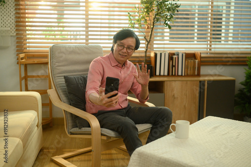 Mature Asian man sitting comfortably at home, smiling and waving during a video call on his smartphone in a warm living room