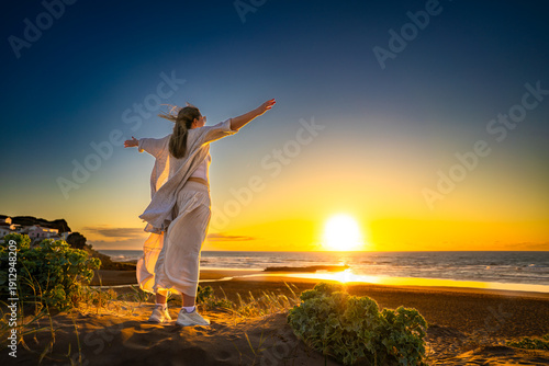 Beautiful middle-aged woman standing with outstretched arms on sandy beach on sunset on windy spring day. Back view. Monte Clerigo beach on Algarve coast in Portugal