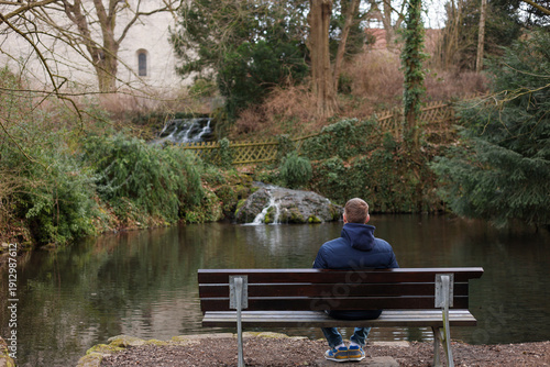 Fotografie A man sits alone on a wooden bench facing a calm pond surrounded by trees and greenery