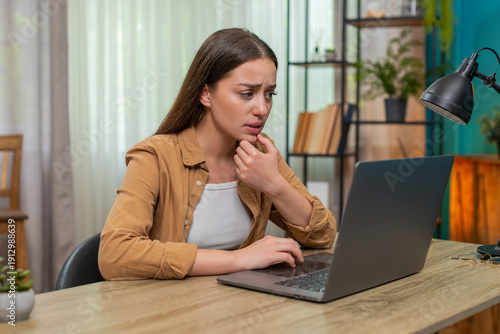 Young woman sitting at home table reads sad news on laptop frowning and showing stress and fatigue. Caucasian girl feels tired and uneasy seeking comfort and hoping for better times ahead tonight now