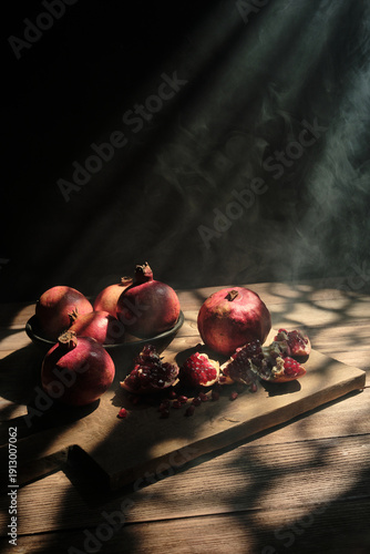 An artistic food composition featuring whole and split pomegranates on a rustic board, illuminated by mystical volumetric light beams with subtle smoke or steam swirls in a dark room