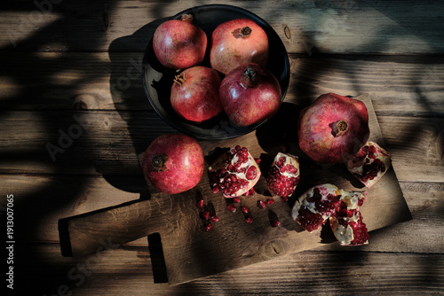 Flat lay photography of fresh pomegranates in a bowl and broken pieces with red seeds on a rustic cutting board, featuring artistic natural shadows on a wooden background.