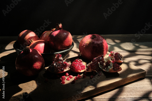 A beautiful food photography scene featuring whole pomegranates and open fruit with glistening seeds on a wooden surface, decorated by natural patterned shadows and warm sunlight