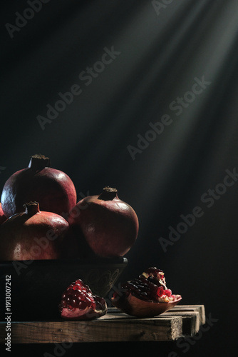 A vertical professional shot of pomegranates in a bowl and sliced pieces on a rustic wood surface, featuring ethereal light beams and smoke swirls against a dark moody background.