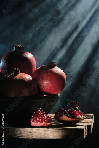 A vertical professional shot of pomegranates in a bowl and sliced pieces on a rustic wood surface, featuring ethereal light beams and smoke swirls against a dark moody background.