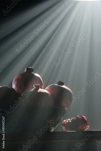 A vertical professional shot of pomegranates in a bowl and sliced pieces on a rustic wood surface, featuring ethereal light beams and smoke swirls against a dark moody background.